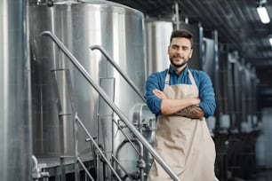 Brewer with large metal container at brewery factory. Smiling young confident attractive guy worker or owner with tattoos in apron, with crossed arms, near metal cauldron in plant interior, free space