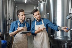 Plant management, modern technology and work at brewery. Check and control equipment. Millennial attractive men workers in aprons, use tablet and operate kettle at plant interior, copy space