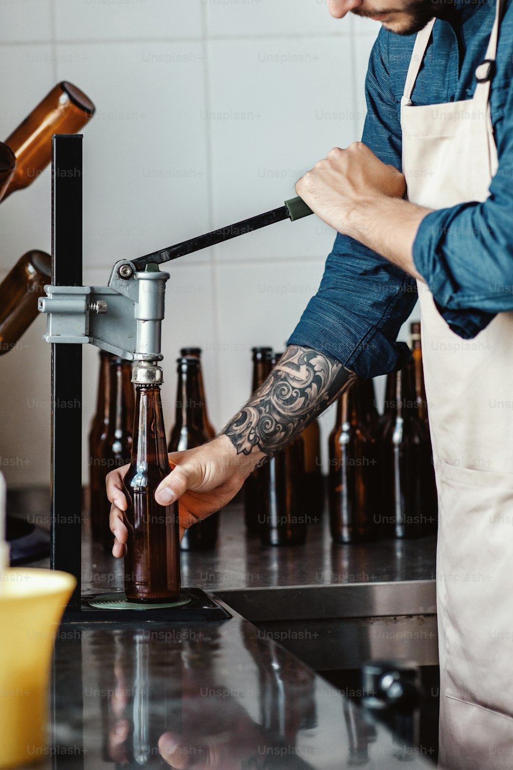 Young man work at beer plant. Brewer with industrial equipment at brewery. Millennial strong guy worker in apron, corks bottles with beer on conveyor belt indoor, vertical, cropped, free space