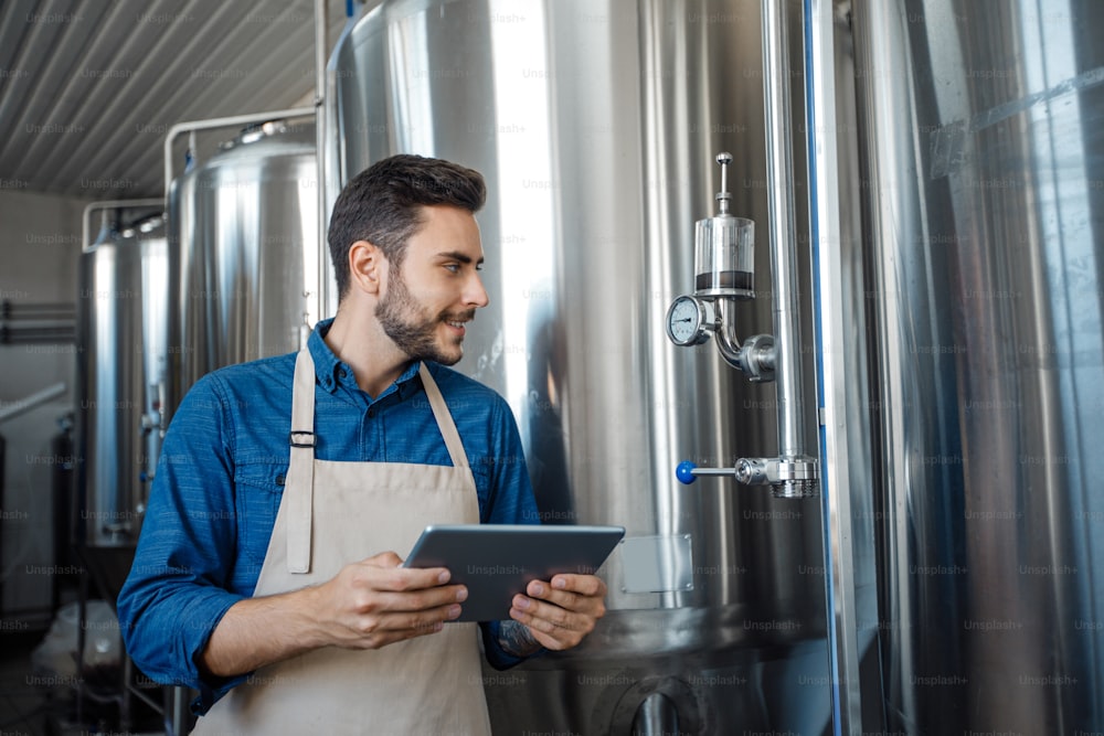 Brewery, modern business and craft beverage production management with device. Cheerful young handsome man in apron with tablet, control equipment, looks at large metal tank with craft drink