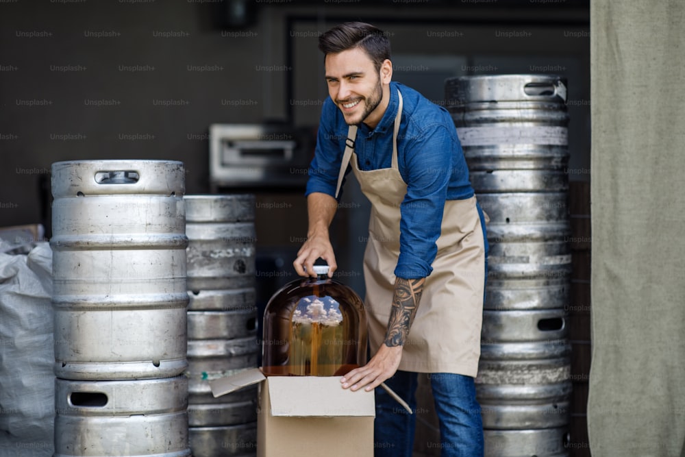 Small business, factory job and startup during covid. Brewery, beer production, equipment and ad of alcohol. Smiling young handsome guy brewer working in warehouse with metal barrels, empty space