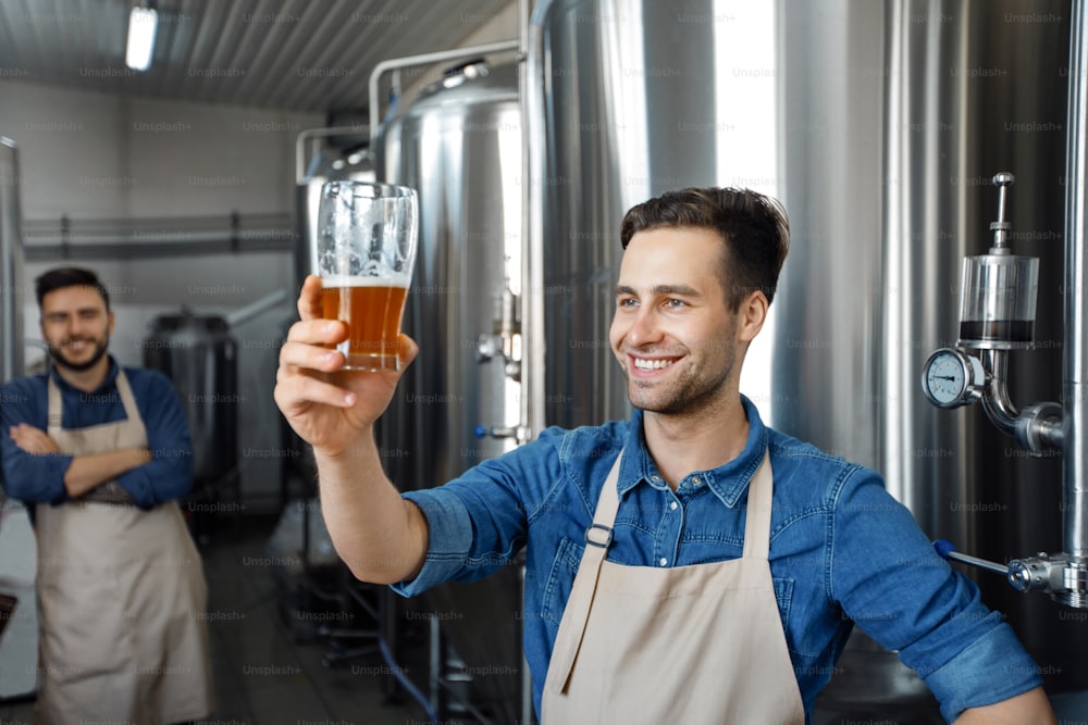 Family small business. Professional brewer on own craft alcohol production. Happy millennial proud handsome man in apron check quality of beer near large metal tank with craft drink, empty space