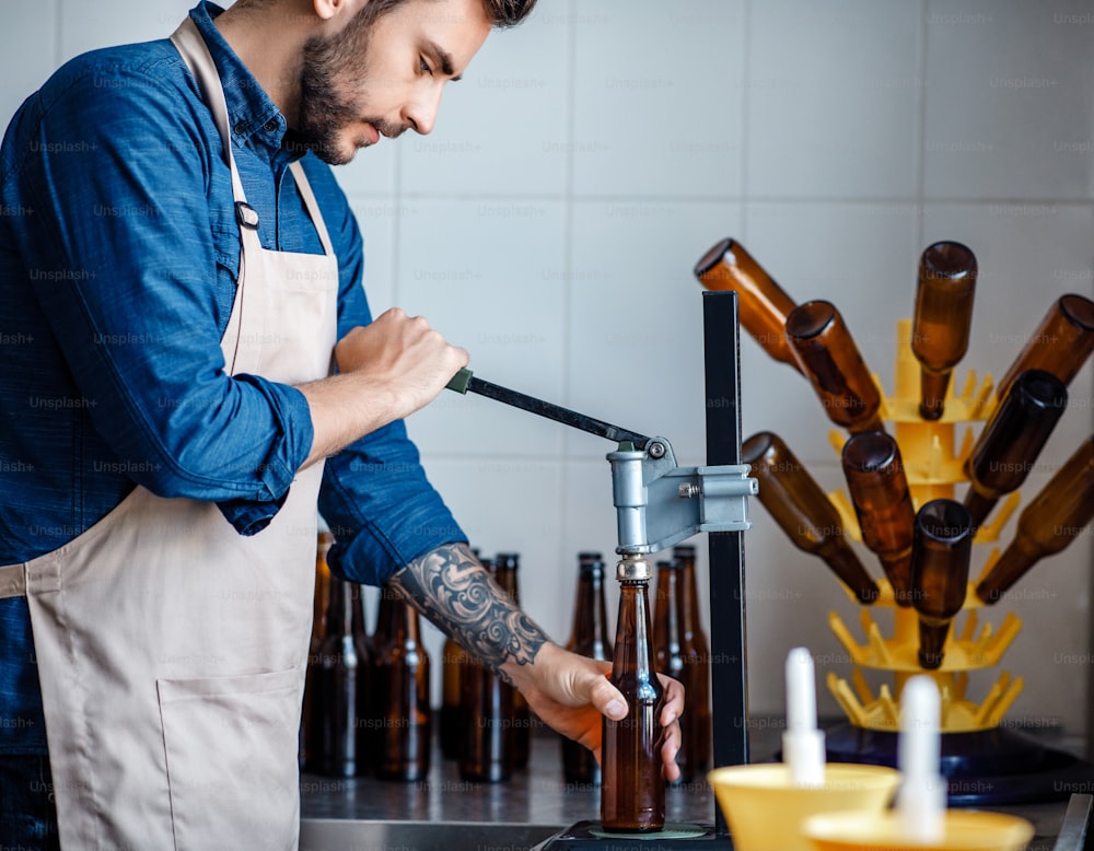 Small business, startup and brewery work. Craft drink production at modern plant. Concentrated young attractive bearded man in apron corks beer bottles on conveyor belt in factory, empty space