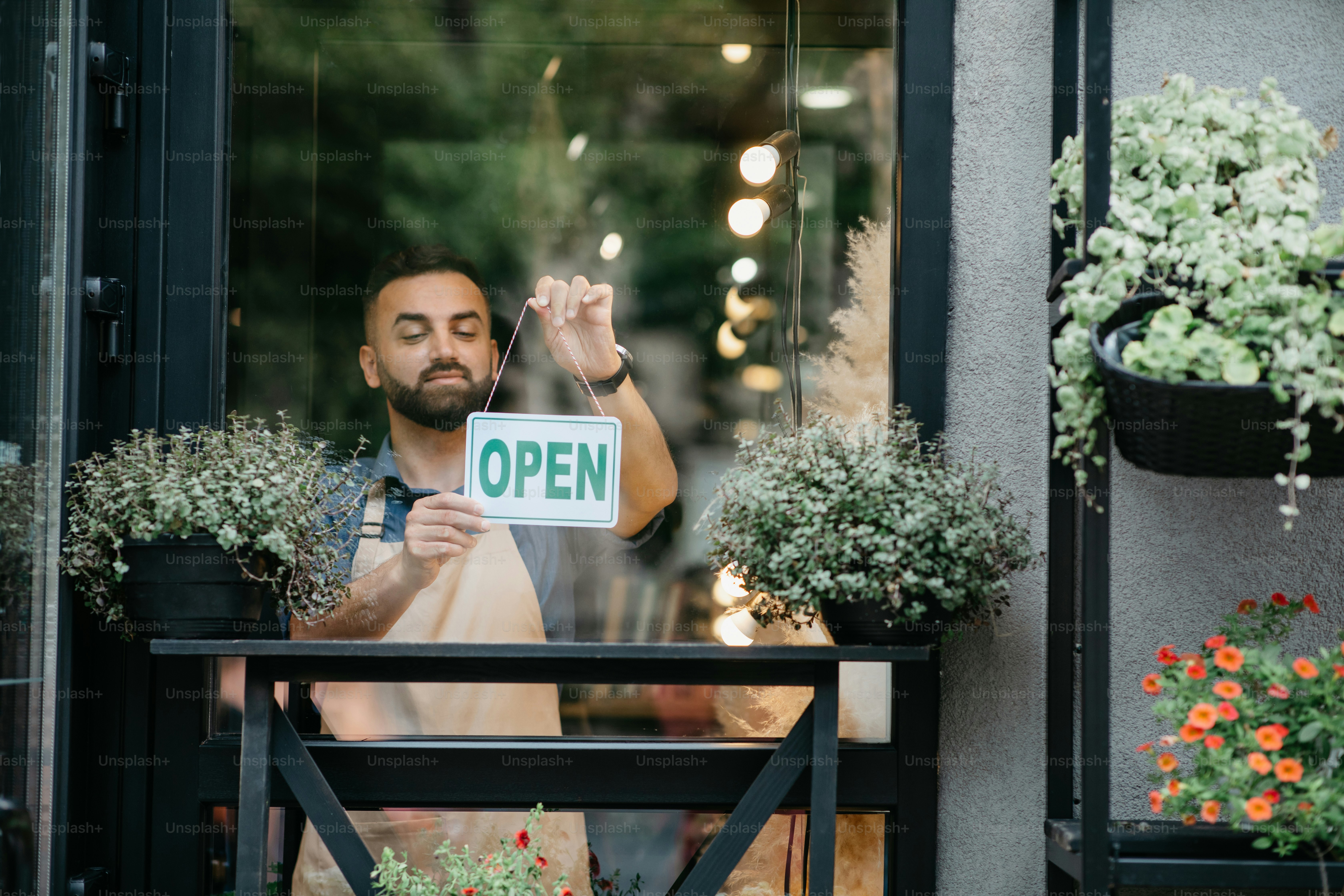 Business concept, caucasian man holding open sign indoors in shop. Young man flower center employee in apron standing in garden center. Millennial guy in studio with plants in pots turns nameplate