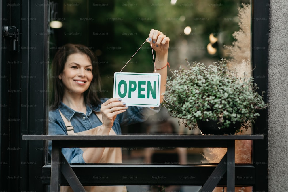 We are working again, job concept and opening flower shop after covid-19 lockdown. Cheerful millennial female in apron turns sign with inscription open in plants studio with fresh bouquet in morning