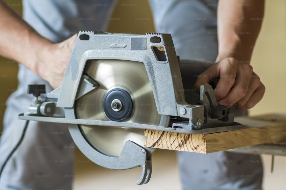 Close-up of muscular carpenter hands using new shiny modern powerful circular sharp electrical saw for cutting hard wooden board. Professional tools for construction and building concept.