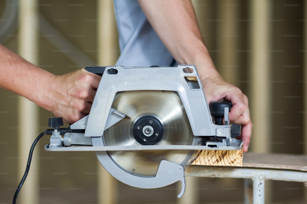 Close-up of muscular carpenter hands using new shiny modern powerful circular sharp electrical saw for cutting hard wooden board. Professional tools for construction and building concept.