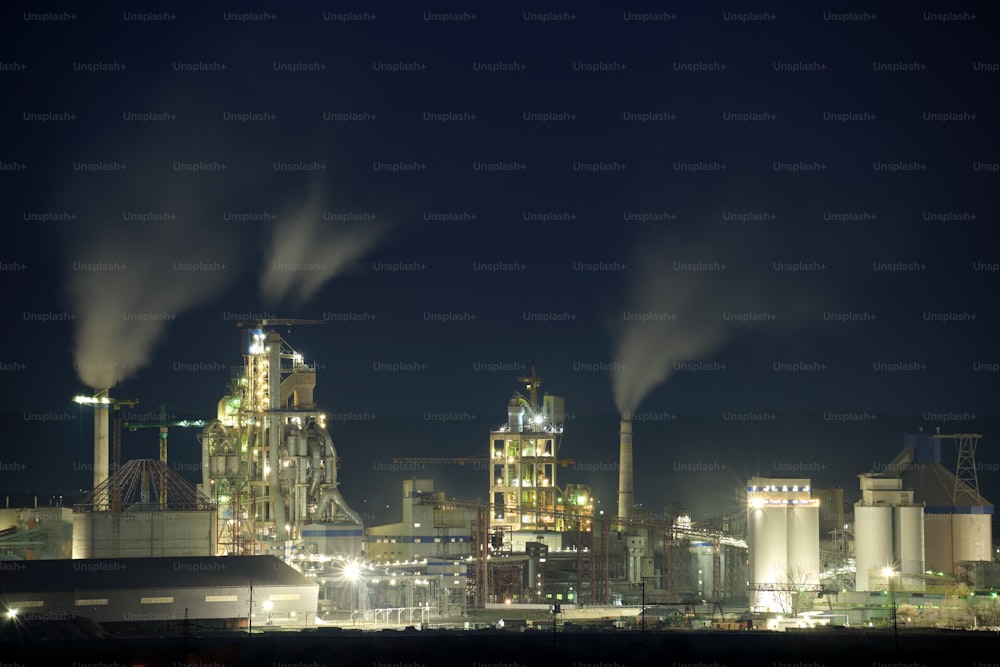 Planta de cemento iluminada con alta estructura de fábrica y grúas torre en el área de producción industrial por la noche. Fabricación y concepto de industria global.