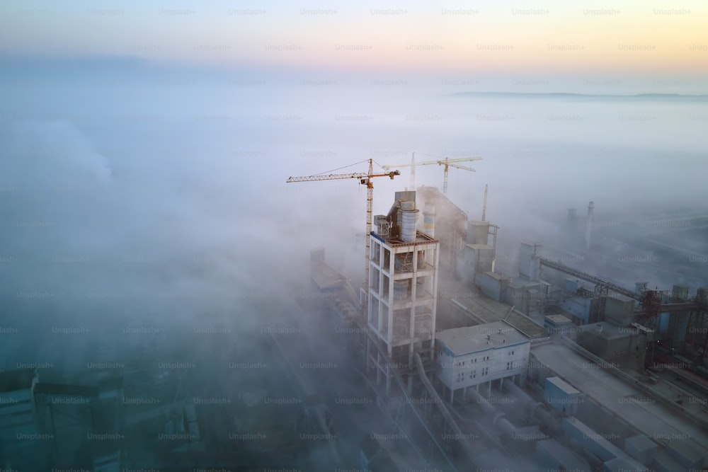 Vista aerea della fabbrica di cemento con alta struttura dell'impianto in calcestruzzo e gru a torre nel sito di produzione industriale nella sera nebbiosa. Produzione e concetto di industria globale.