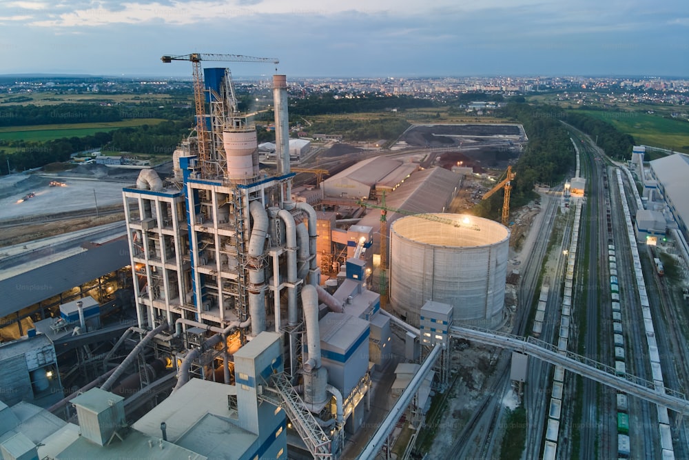 Vista aerea di cementificio con alta struttura impiantistica in calcestruzzo e gru a torre nell'area di produzione industriale di notte. Produzione e concetto di industria globale.