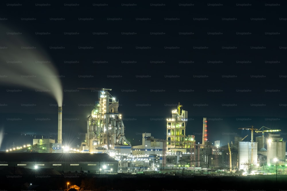 Haute structure en béton de l’usine de ciment avec grue à tour et cheminée fumante la nuit. Production industrielle et concept de pollution atmosphérique.