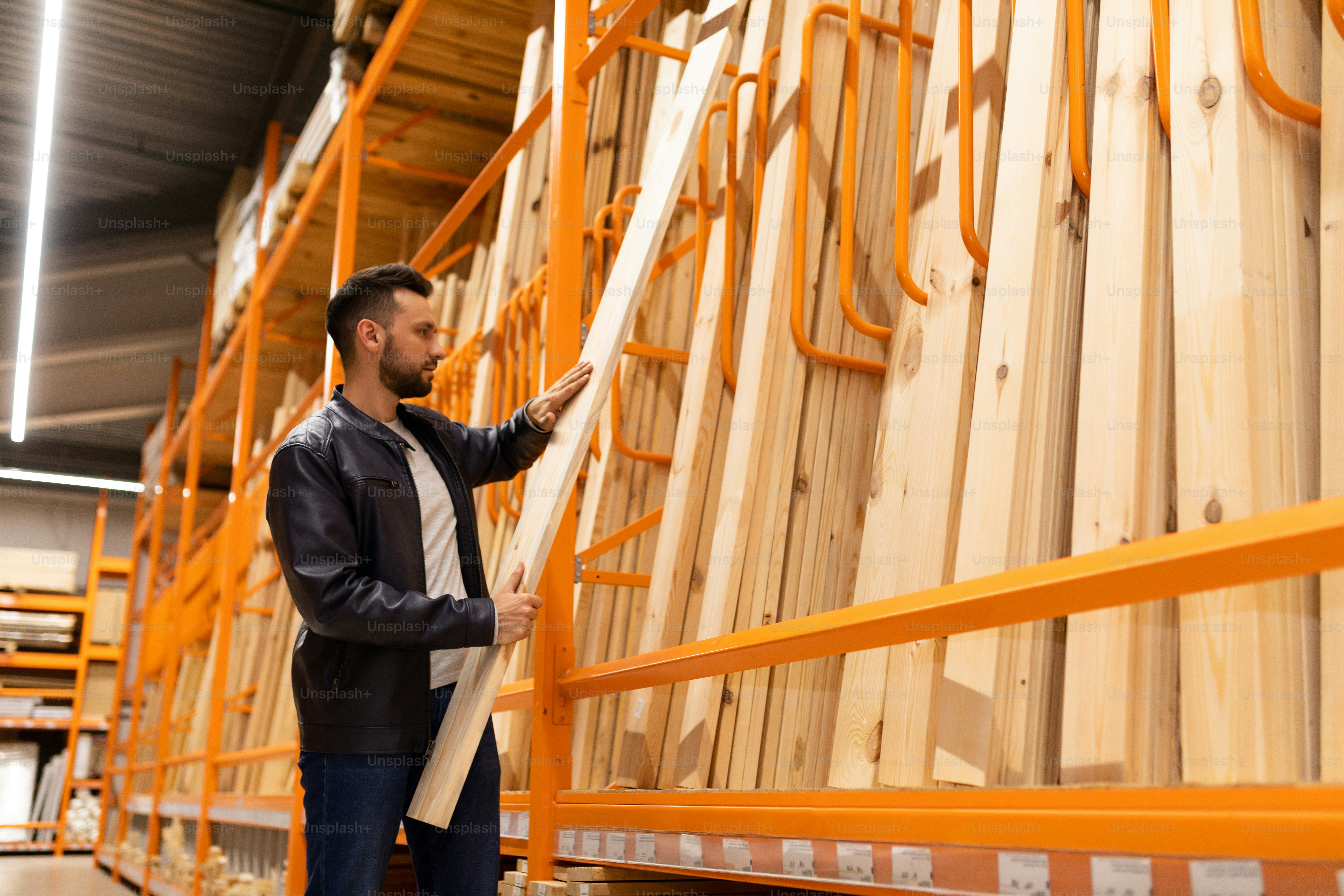 Un homme dans un hypermarché de la construction du département du bois tient une planche dans ses mains à côté d’un support avec des barres de bois et des planches à clins pour la décoration des murs et des façades.