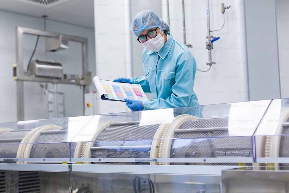 Kaukasischer Fabrikarbeiter im blauen Laboranzug, mit Tablet in den Händen, beugt sich auf der Herstellungsmaschine, überprüft die Messwerte, schaut in die Kamera