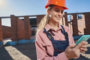 Waist-up photo of a pleasant lady in hardhat and overalls checking her phone and smiling