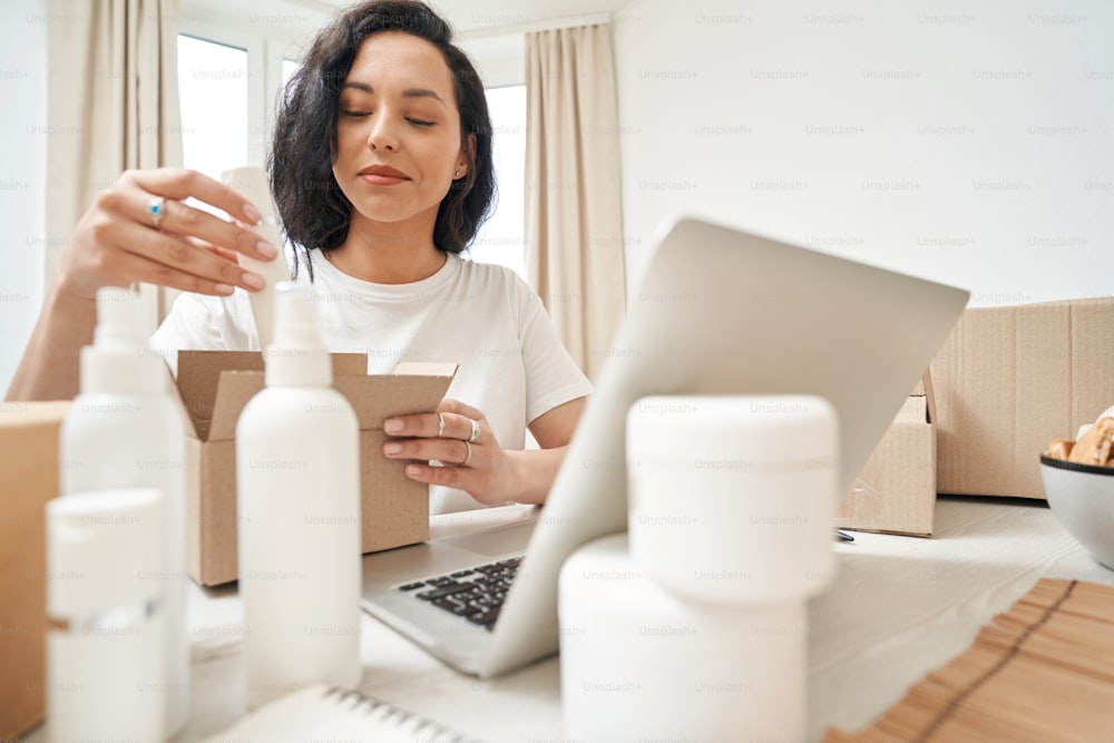 Focused lady sitting at laptop and putting beauty product into cardboard box