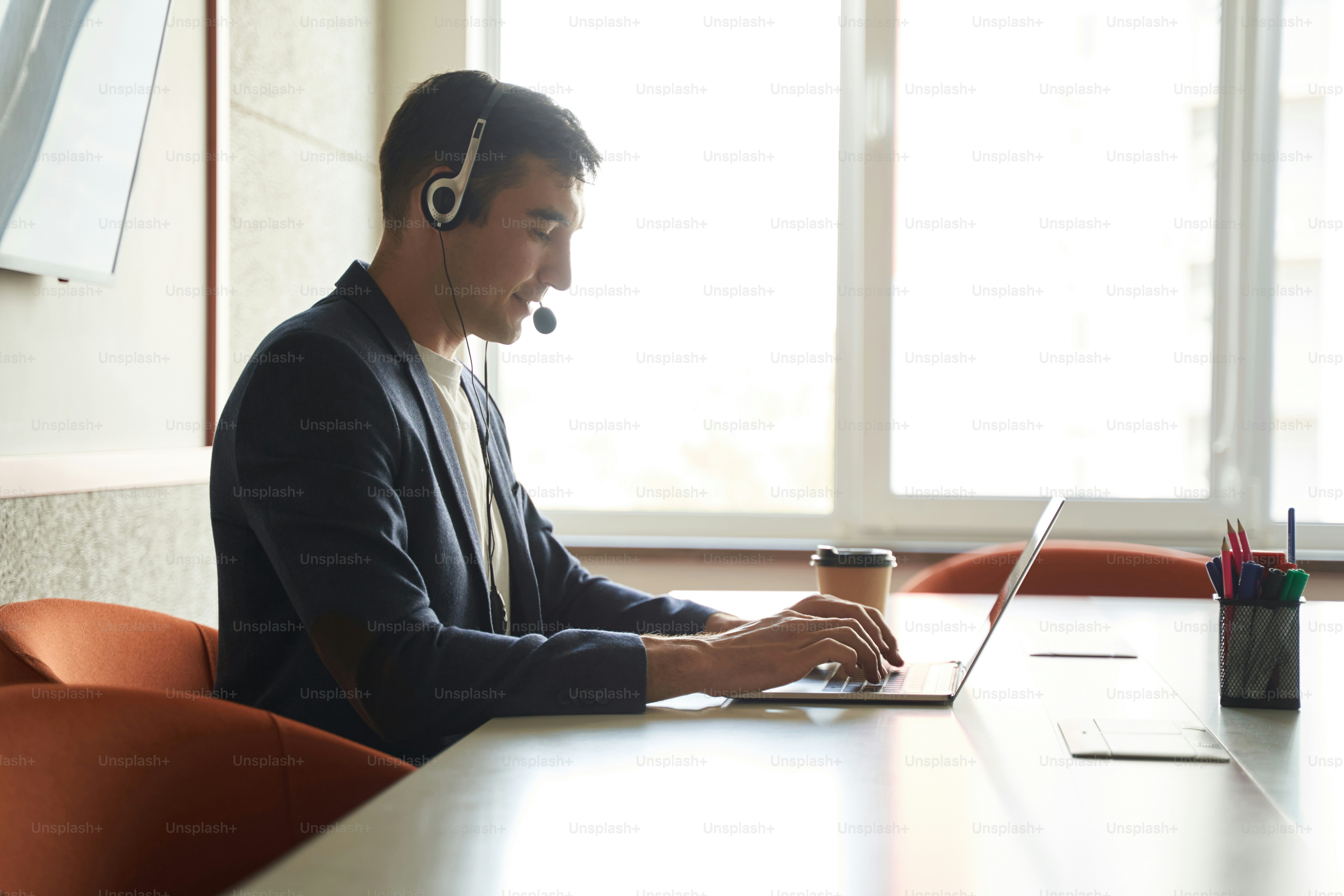 Male working online with help of laptop and headset while texting on keyboard and speaking into microphone