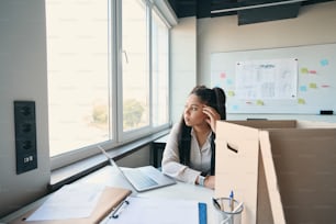 Pensive online shop employee seated at office desk with cardboard box looking out of window