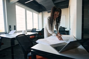 Focused female architect leaning over rolled-out technical drawing while looking at laptop screen