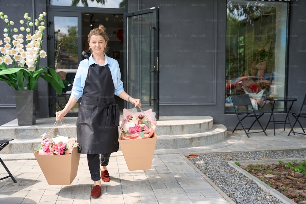 Female florist in apron carries huge package with luxurious bouquets of flowers for delivery to buyer