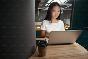 Retrato de cintura para arriba de un feliz y alegre oficinista asiático está sentado en el escritorio y leyendo algo en la computadora en una oficina de espacio abierto