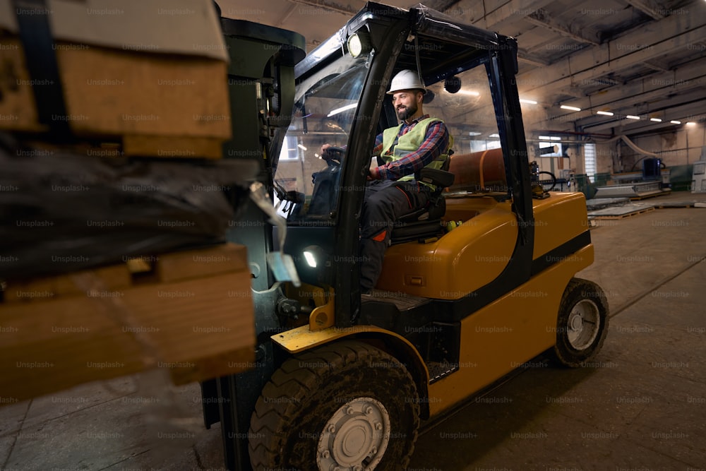 Man in protective clothes and helmet sitting in the loading machine and driving in manufactory