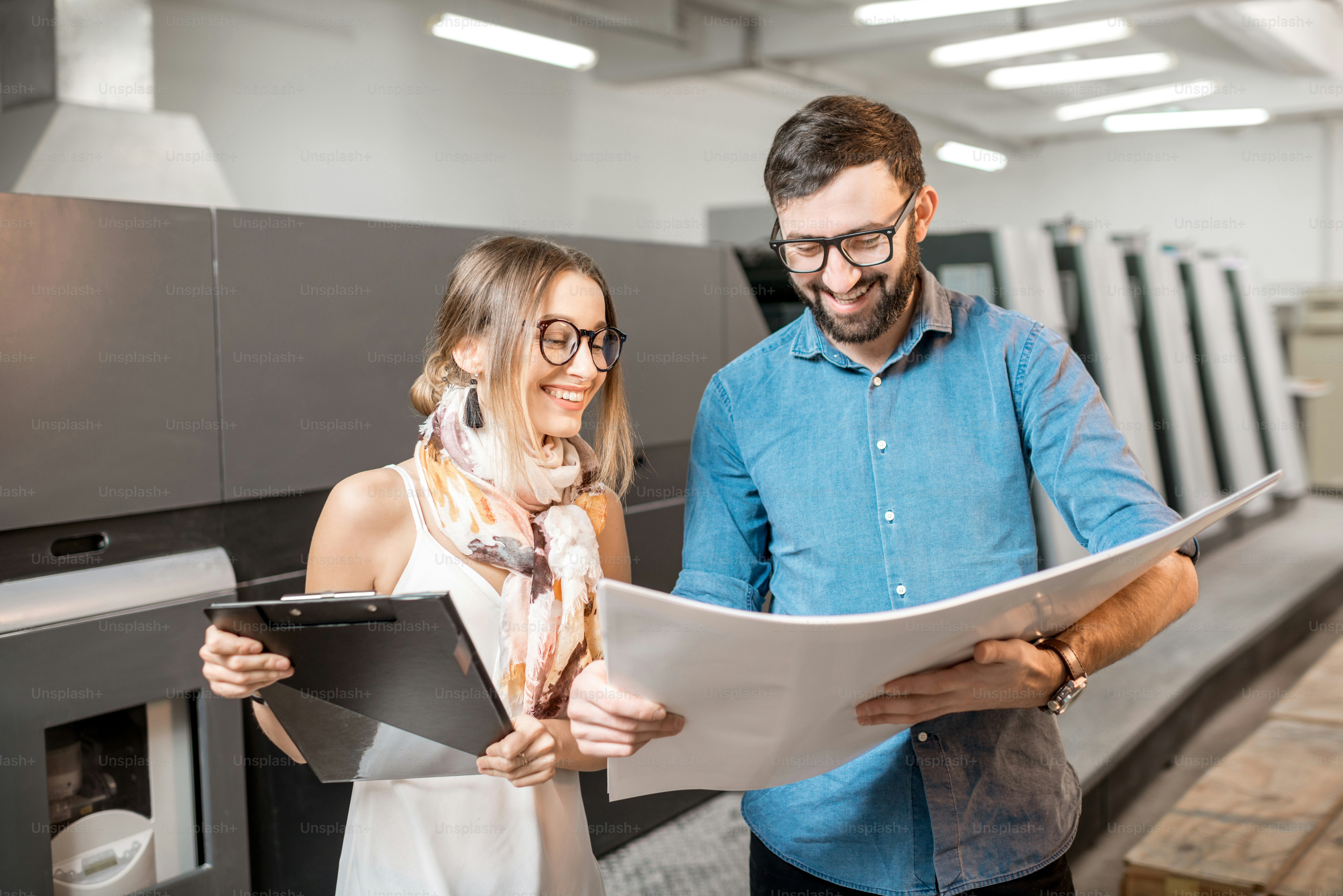印刷物製造で立つ紙の印刷物と一緒に働く若い女性デザイナーと版画オペレーター