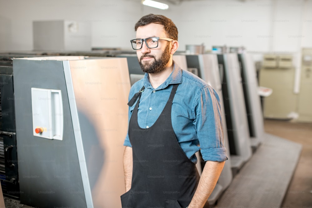 Ritratto di un bel tipografo in piedi alla produzione di stampa con macchina offset sullo sfondo
