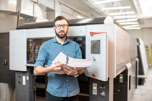 Portrait d’un beau typographe debout avec une pile de papier à l’imprimerie avec une machine offset en arrière-plan