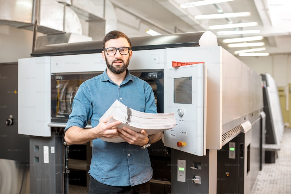 Ritratto di un bel tipografo in piedi con pila di carta alla produzione di stampa con macchina offset sullo sfondo