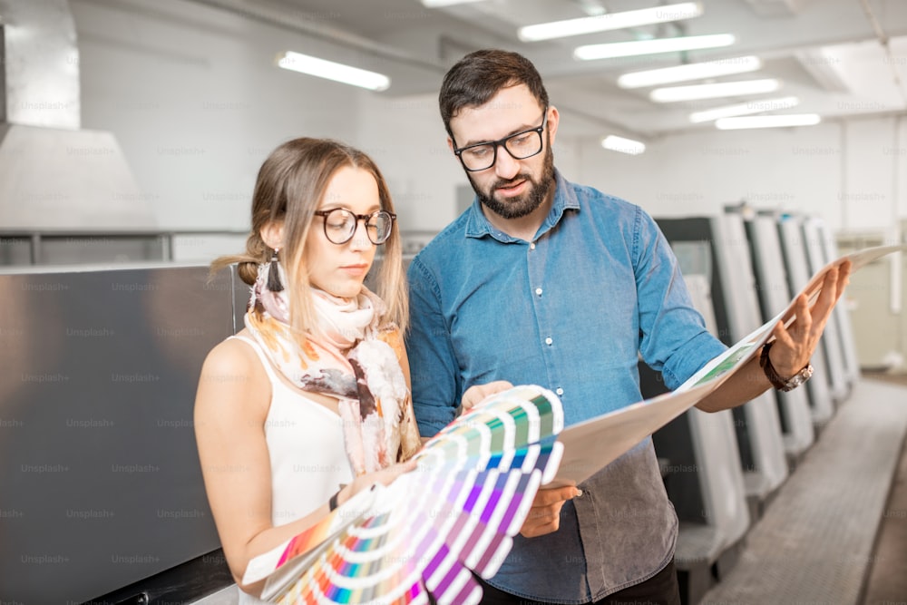 젊은 여성 디자이너와 인쇄 운영자가 인쇄 제조소에 서 있는 색상 견본과 함께 일하고 있다