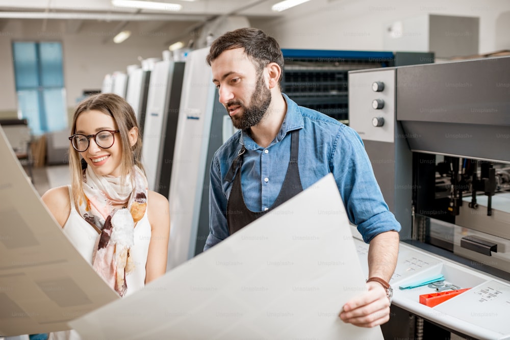 Young woman designer and print operator working with paper print at the print manufacturing with offset machine on the background