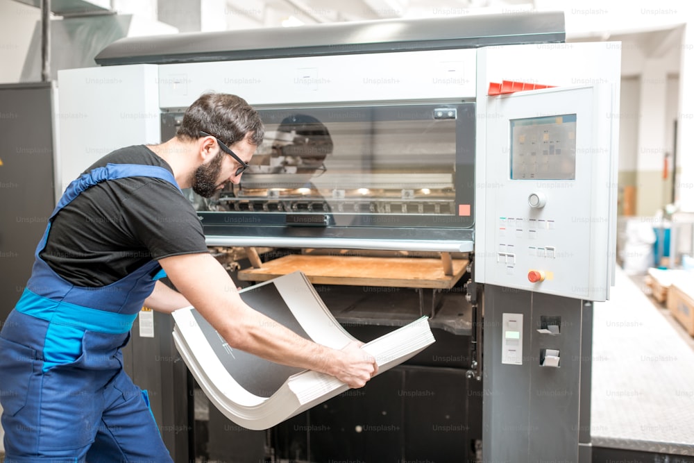 Worker in unifrom getting printed sheets from the offset machine at the printing manufacturing