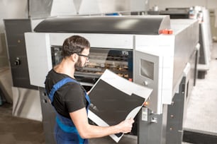 Tipógrafo verificando a qualidade de impressão em pé perto da máquina offset na fabricação de impressão