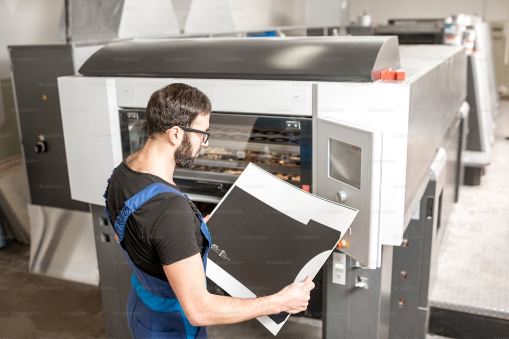 Tipografo che controlla la qualità di stampa in piedi vicino alla macchina offset durante la produzione di stampa