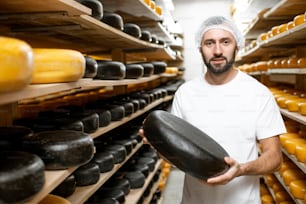 Trabalhador segurando roda de queijo coberta com cera preta no armazenamento com prateleiras cheias de queijo durante o processo de envelhecimento