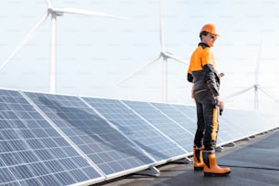Lavoratore ben attrezzato in abbigliamento protettivo arancione che esamina i pannelli solari su un impianto fotovoltaico sul tetto. Concetto di manutenzione e installazione di stazioni solari