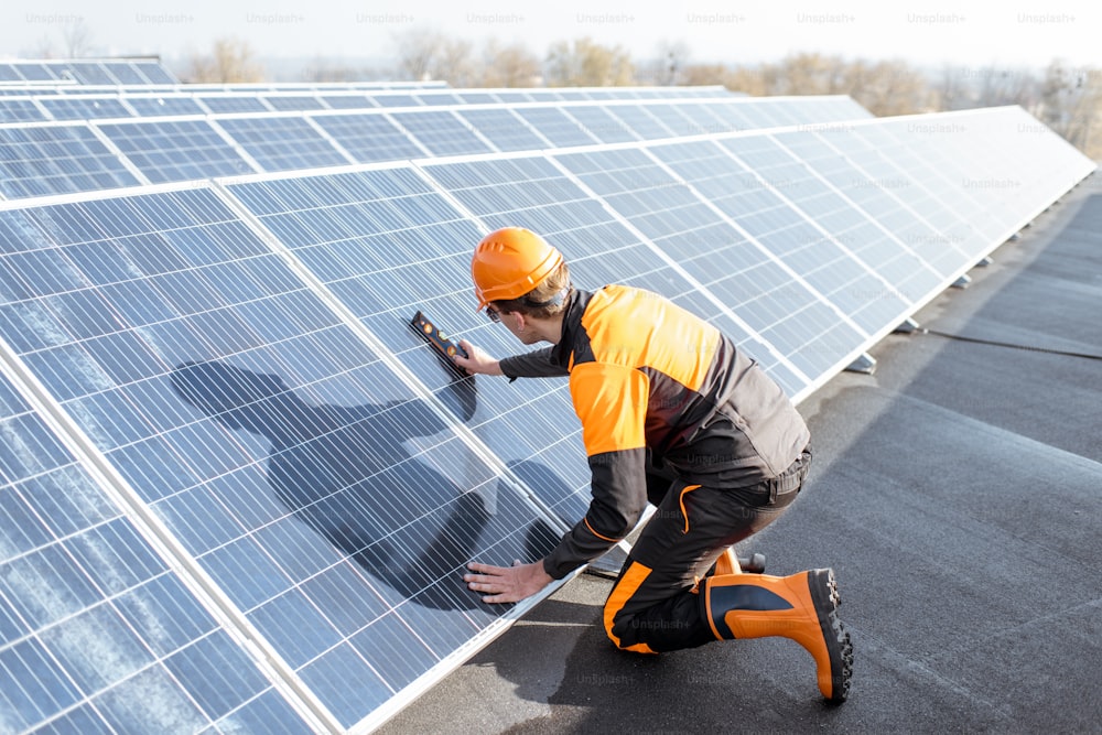 Gut ausgestatteter Arbeiter in orangefarbener Schutzkleidung, der Sonnenkollektoren installiert und den Neigungswinkel auf einer Photovoltaik-Dachanlage misst