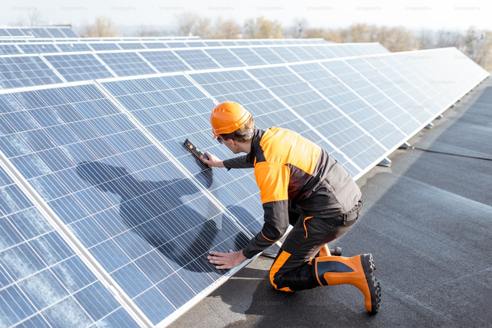 Travailleur bien équipé en vêtements orange de protection installant des panneaux solaires, mesurant l’angle d’inclinaison sur une centrale photovoltaïque sur toit