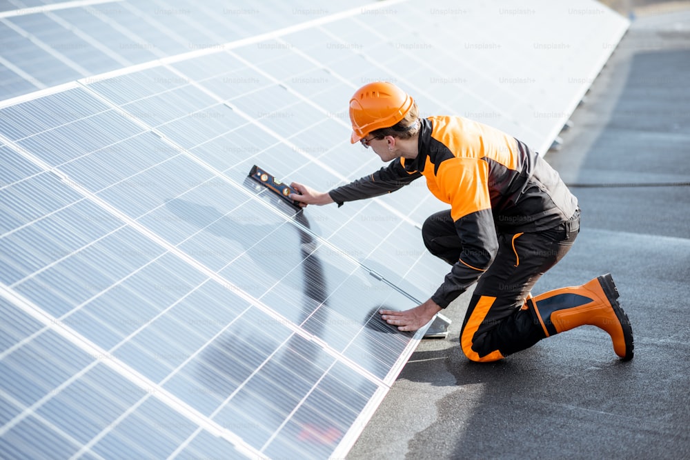 Travailleur bien équipé en vêtements orange de protection installant des panneaux solaires, mesurant l’angle d’inclinaison sur une centrale photovoltaïque sur toit