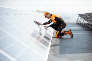 Trabajador bien equipado con ropa protectora naranja instalando paneles solares, midiendo el ángulo de inclinación en una planta fotovoltaica en la azotea