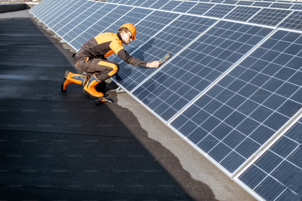 Trabajador bien equipado con ropa protectora naranja instalando paneles solares, midiendo el ángulo de inclinación en una planta fotovoltaica en la azotea