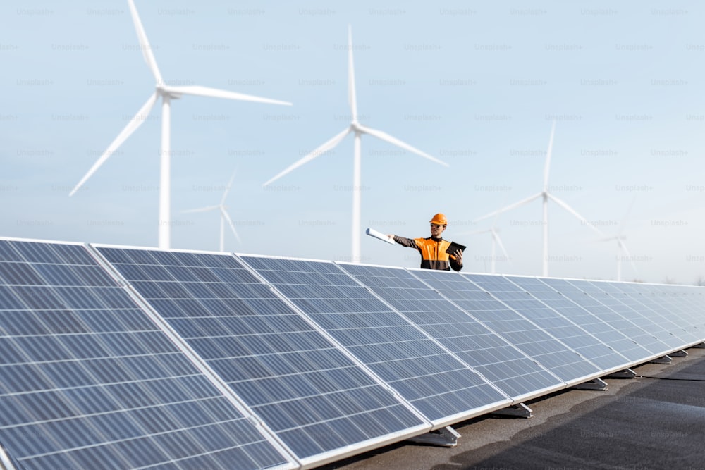 Vista de la planta de energía solar en la azotea con un ingeniero en ropa de trabajo protectora y turbinas eólicas en el fondo. Concepto de energía alternativa y su mantenimiento