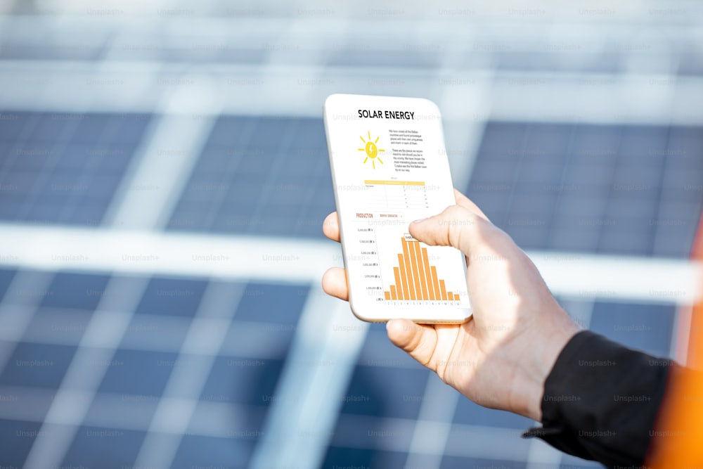 Man examining genaration of solar power plant, holding a smart phone with chart of electricity production. Concept of online monitoring of the electric station