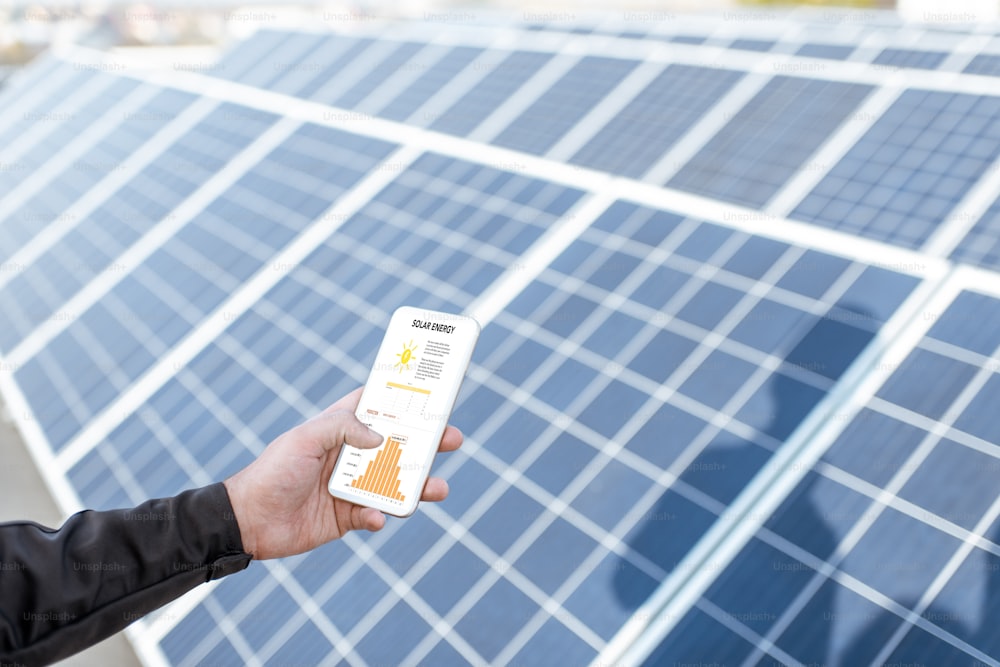 Ein Mann, der die Entstehung eines Solarkraftwerks untersucht, hält ein Smartphone mit einem Diagramm der Stromerzeugung in der Hand. Konzept der Online-Überwachung der Elektrizitätsstation