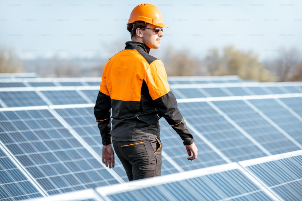 Trabalhador bem equipado em roupas laranja protetoras examinando painéis solares em uma usina fotovoltaica no telhado. Conceito de manutenção e instalação de estações solares