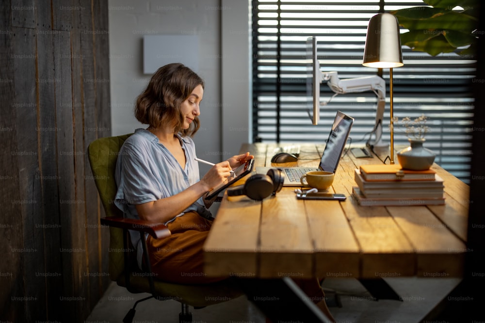 Giovane donna vestita casualmente con un po 'di lavoro creativo, disegnando su una tavoletta digitale, seduta nell'accogliente ed elegante ufficio di casa