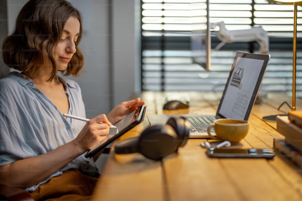 Young woman dressed casually having some creative work, drawing on a digital tablet, sitting at the cozy and stylish home office