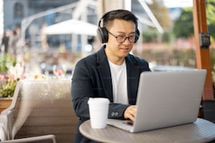 Asian businessman in earphones typing on laptop during work in cafe. Concept of remote and freelance work. Focused adult successful man wearing suit and glasses sitting at table with coffee