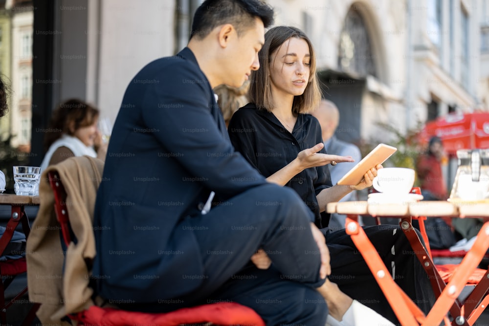 Caucasian businesswoman showing something on digital tablet to asian businessman at outdoor cafe. Concept of remote and freelance work. Idea of teamwork and business cooperation. Sunny day