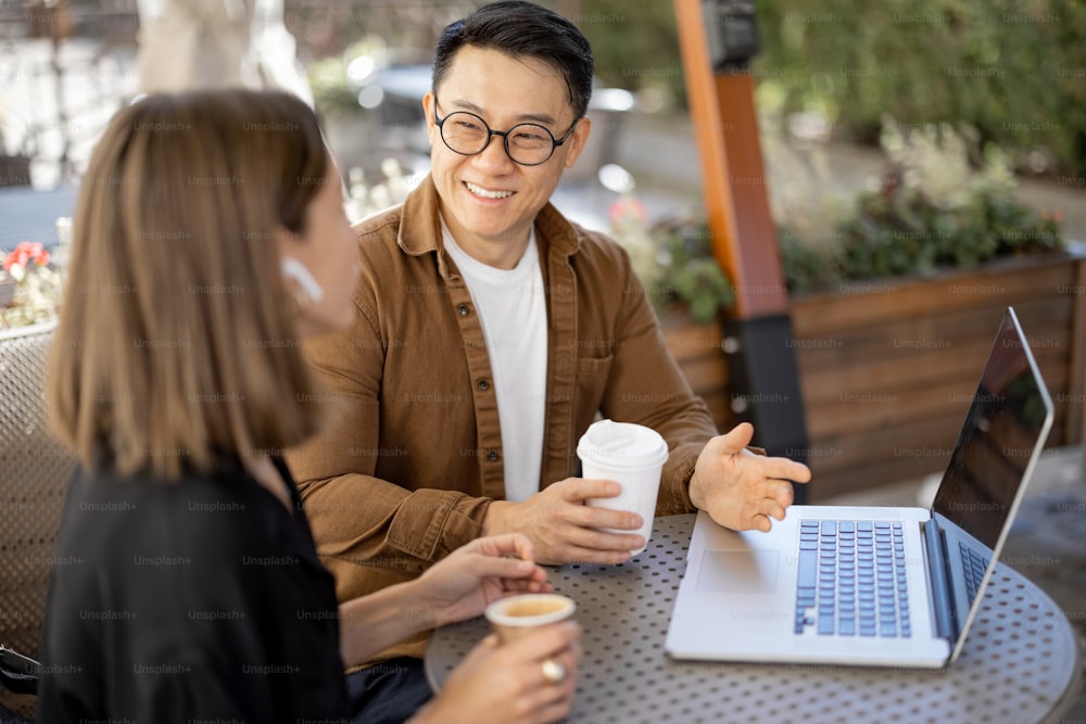 Multiracial business people talking and working with laptop in cafe. Concept of remote and freelance work. Idea of teamwork and business cooperation. Caucasian woman and asian man drinking coffee